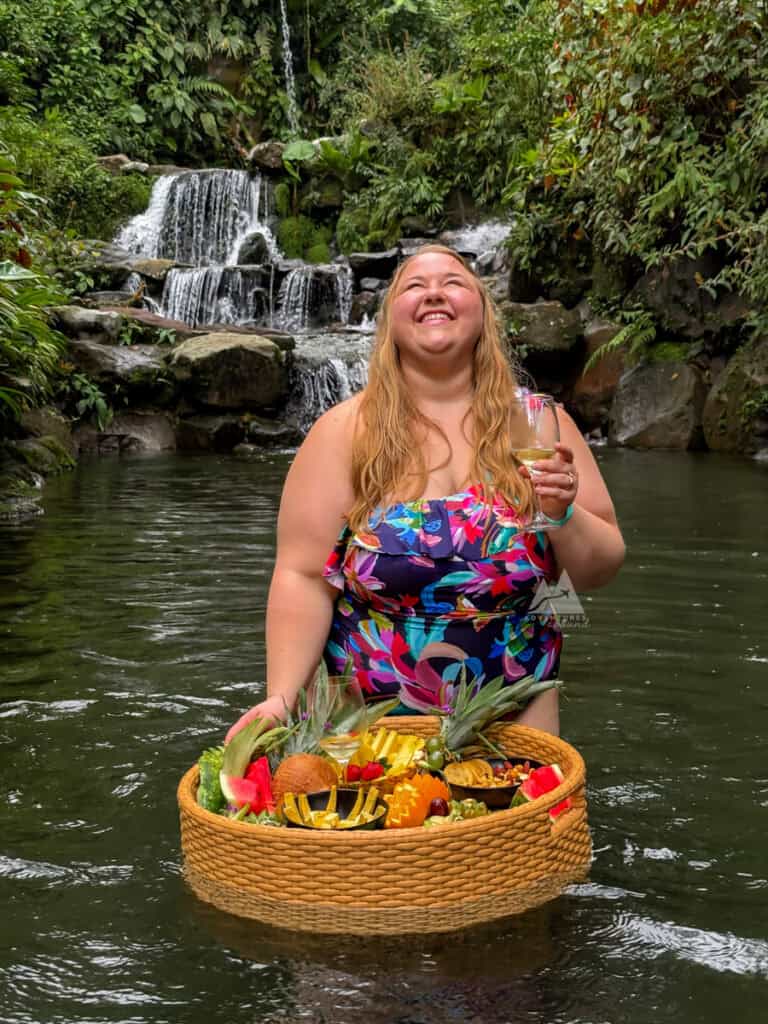 Woman smiling while holding a glass of white wine and a floating tray filled with fresh fruit and cheese in the natural cold spring at Kuru, with a small waterfall cascading behind her. A refreshing and scenic stop near Arenal 1968 in La Fortuna.