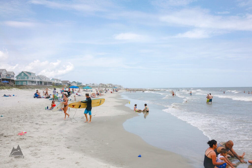 A busy sandy shoreline with people swimming, relaxing, and carrying surfboards along the water at Folly Beach in Charleston. Beach houses line the coast in the background under a hazy blue sky.