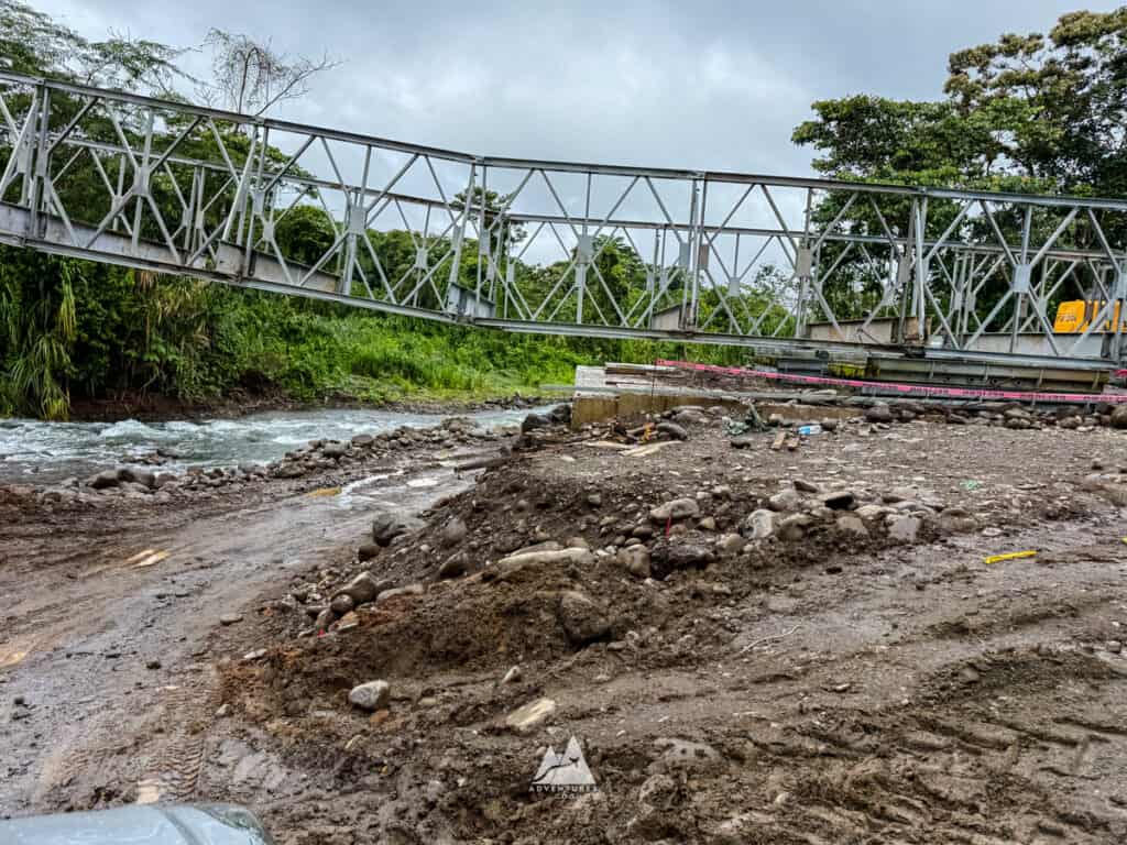 A damaged rural bridge over a fast moving river with muddy, uneven ground and construction debris scattered nearby under a cloudy sky. This highlights potential road and infrastructure challenges to consider when renting a car in Costa Rica.