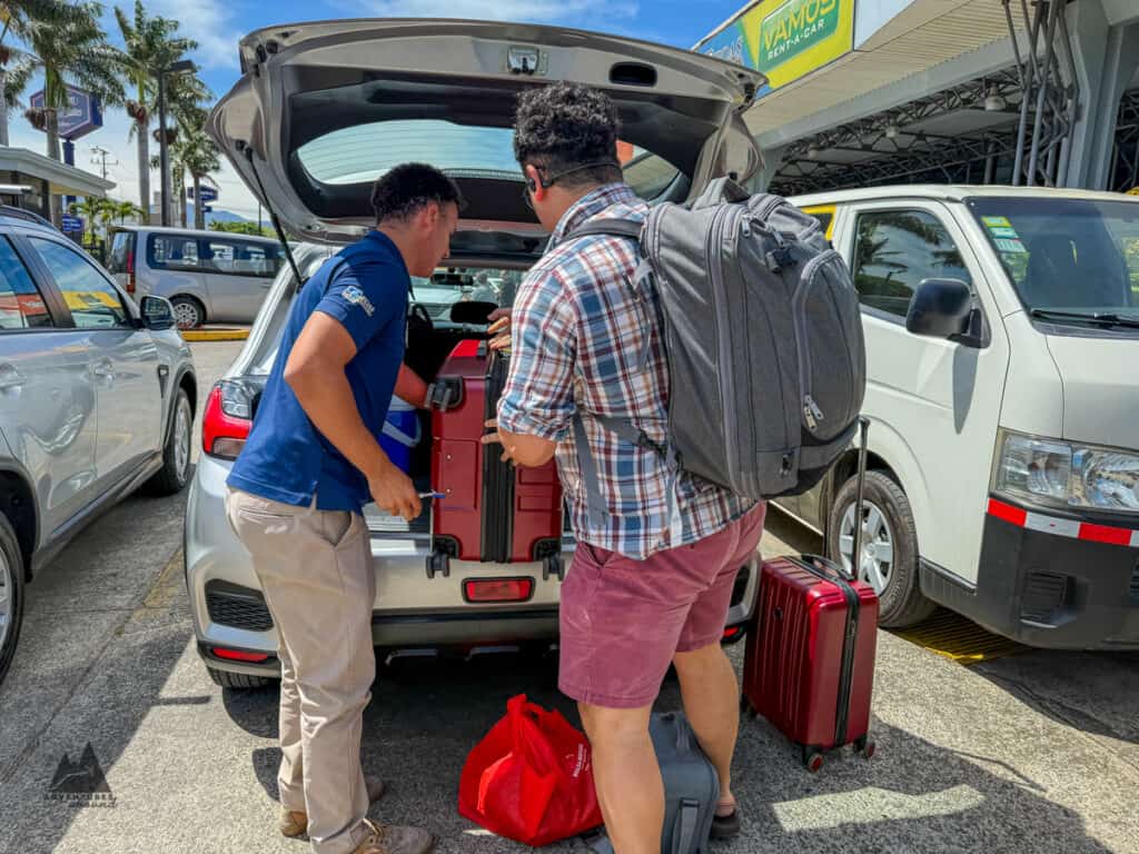 Two men load red suitcases into the trunk of a compact rental car in a sunny parking lot with a sign reading "Vamos Rent A Car" visible above. This moment captures the process of renting a car in Costa Rica and preparing for travel.