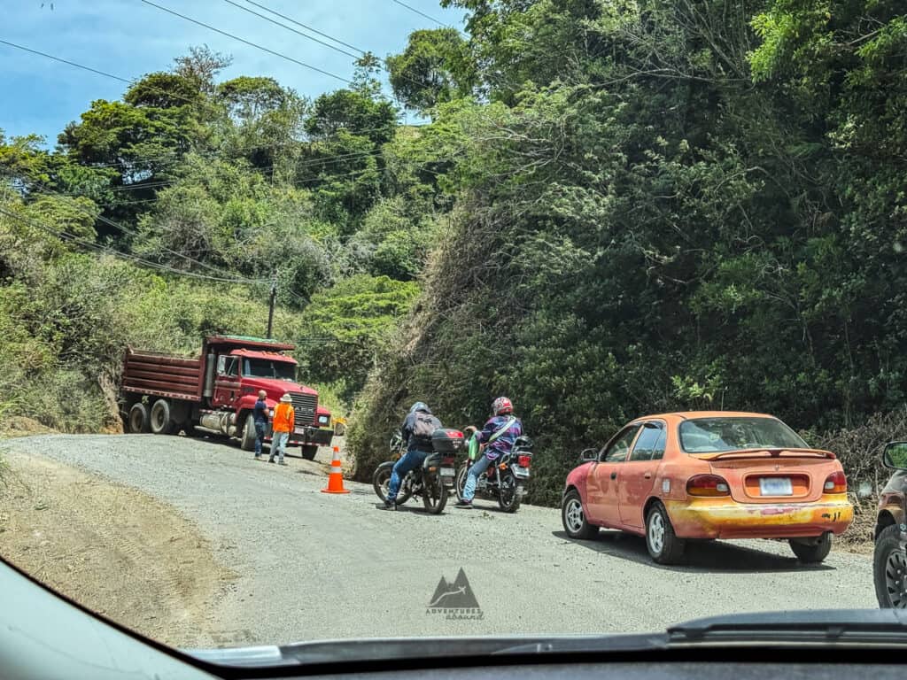 Traffic stopped on a narrow gravel road in Costa Rica as a red dump truck blocks the way and two motorcyclists wait beside a small orange car surrounded by dense green jungle. The scene shows road conditions travelers may encounter when renting a car in Costa Rica.
