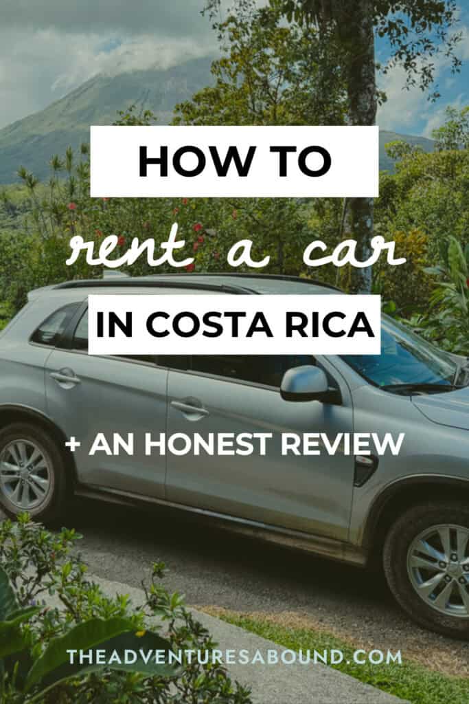 A silver SUV parked along a scenic roadside with tropical greenery and a volcano partially covered by clouds in the background. Text overlay reads "HOW TO rent a car IN COSTA RICA + AN HONEST REVIEW THEADVENTURESABOUND.COM."