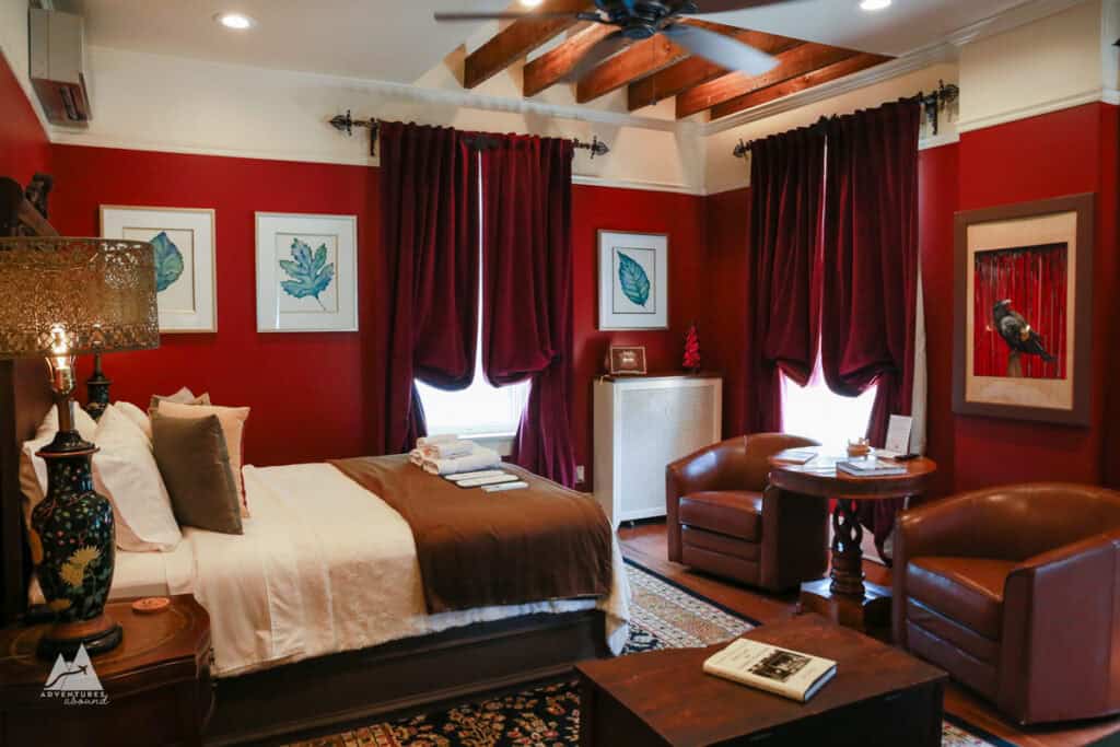 A view of my hotel room at Hygrade Inn: Walls are painted deep red with darker red curtains draped from near the ceiling and art along the walls, a bed to the left with white linens and a brown blanket at the foot of the bed, and two leather chairs on the other edge of the room.