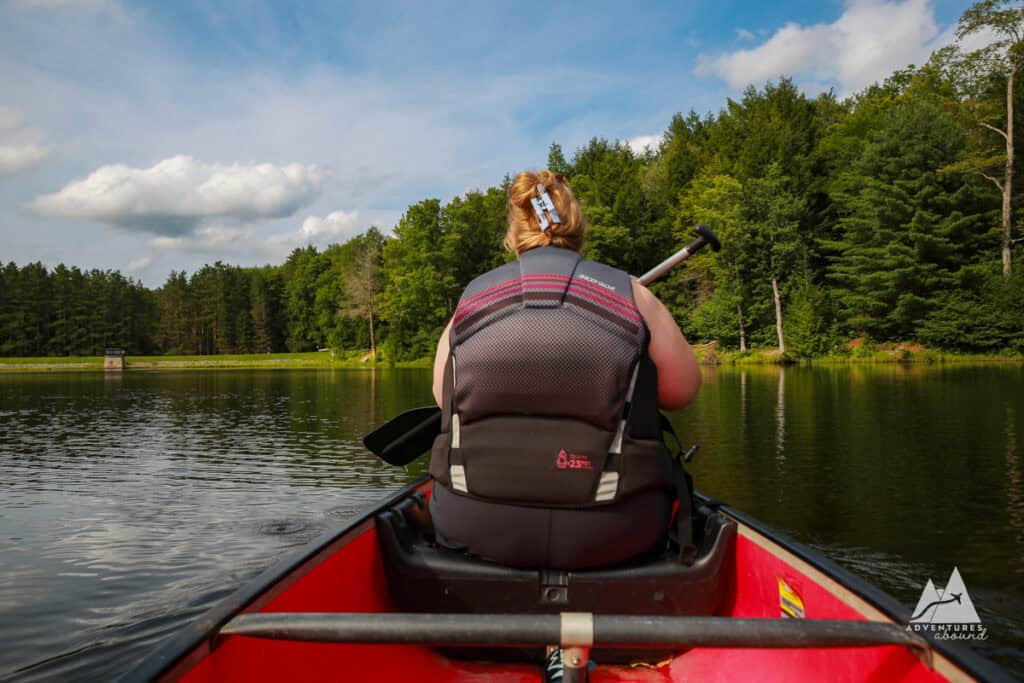 A woman with blonde hair in a clip, wearing a black life jacket, paddles in a red canoe on a serene lake surrounded by lush green pine trees. The scene is peaceful and seems quiet along the pond in Parker Dam State Park.