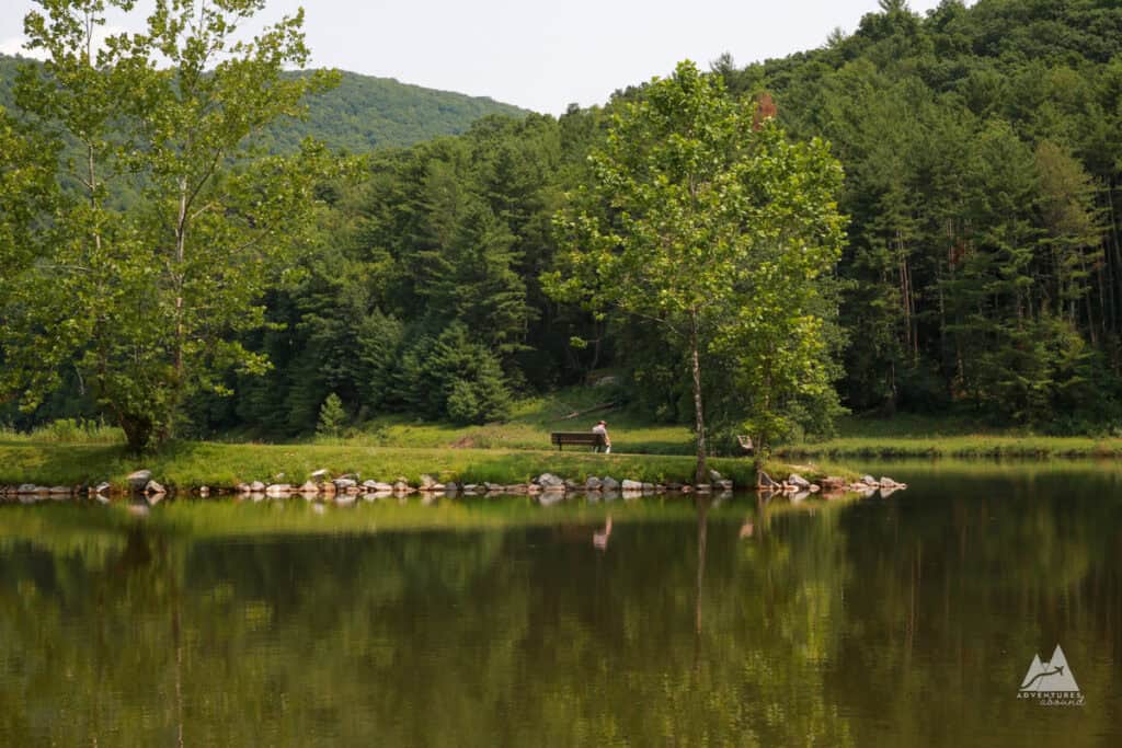 A small strip of land sits along the reservoir, surrounded by deep green trees and lined with stones. A man sits on a bench, tiny in the distance, taking in the beautiful scene at Sinnemahoning State Park.