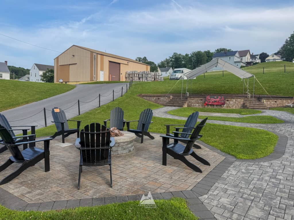 A fire pit made of stone on a stone patio, with black Adirondack chairs surrounding in a circle, with grass on the side. In the distance, a path leads to stone steps and a platform covered with a tent, suggesting a setup for a warm outdoor evening with live music at Straub Brewery/.