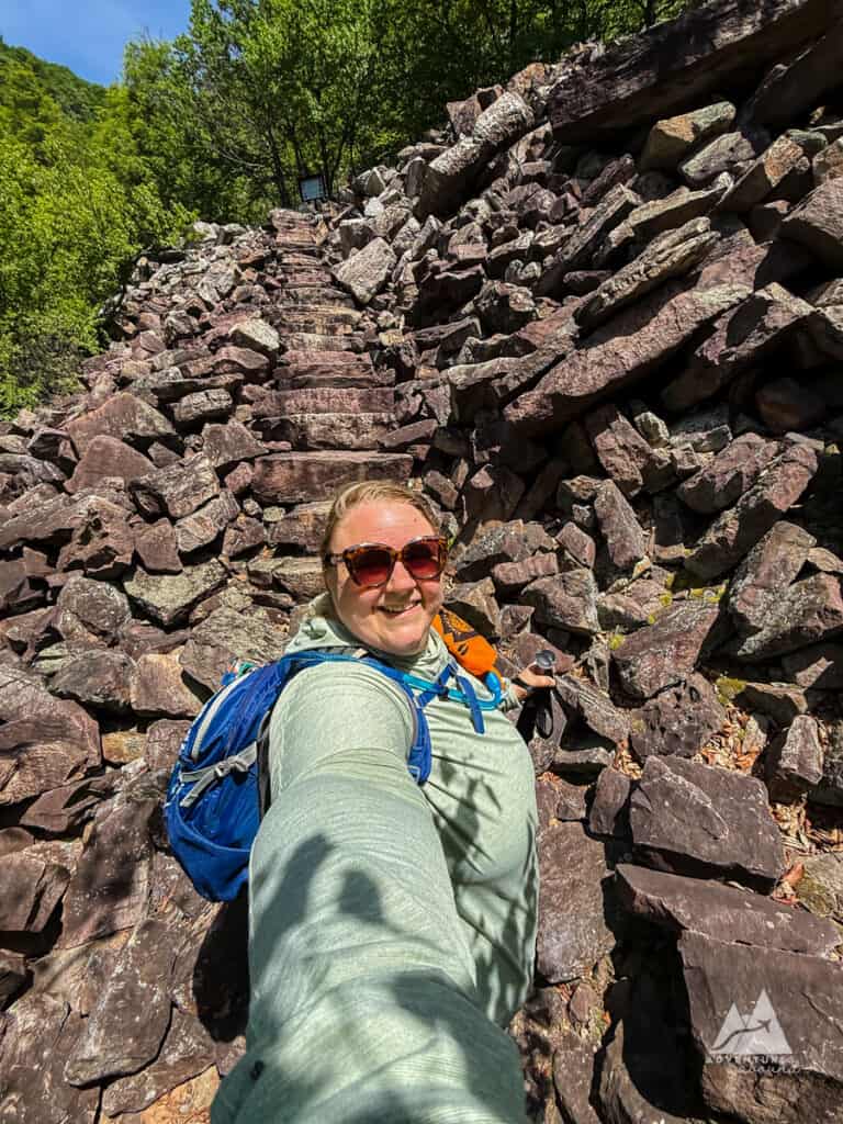 A woman faces the camera, her arm outstretched in a selfie pose, with a blue backpack and sunglasses on. Behind her, a cliff of rocks rises beyond the screen, a matching set of stairs cutting through in a never-ending climb.